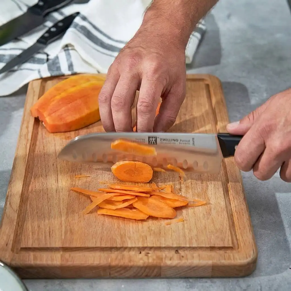 A hand slicing a vibrant orange carrot with a motion-blurred Zwilling Santoku knife on a rustic wooden cutting board under natural window light.